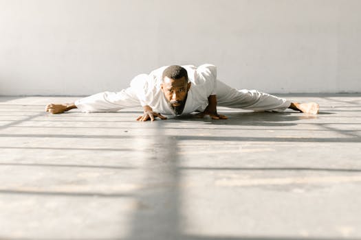 A bearded man in a white outfit performs a floor stretch in a sunlit room, showcasing light and shadow.