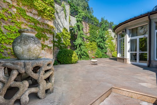Free Exterior of house with large windows placed in spacious yard with aged stone sculptures against high wall covered with green plants in daytime Stock Photo