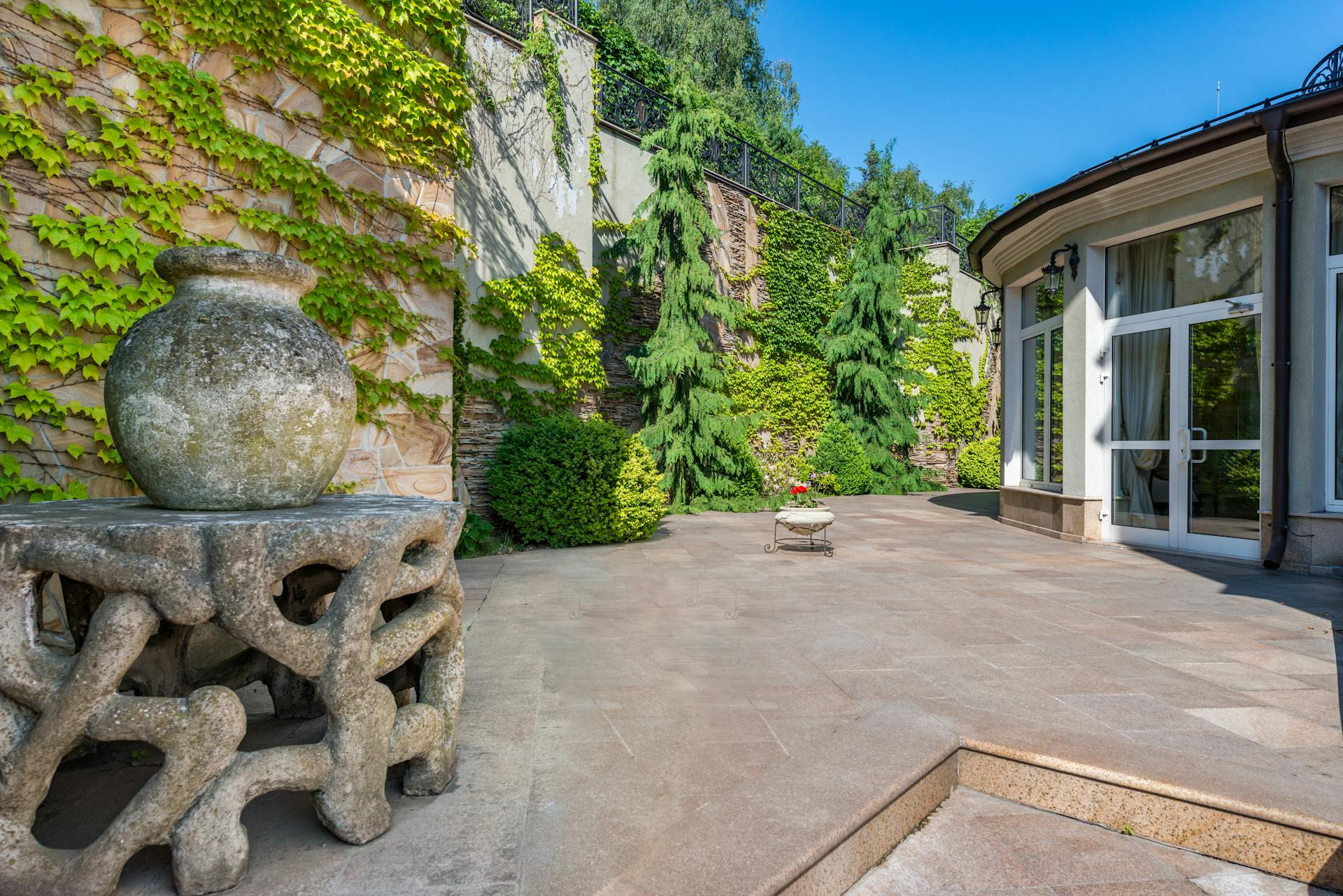 Exterior of house with large windows placed in spacious yard with aged stone sculptures against high wall covered with green plants in daytime