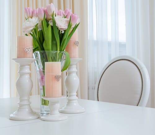 Interior of light dining room with white table with vase with bouquet of flowers and candles in candlesticks near chair next to windows with curtains in daytime