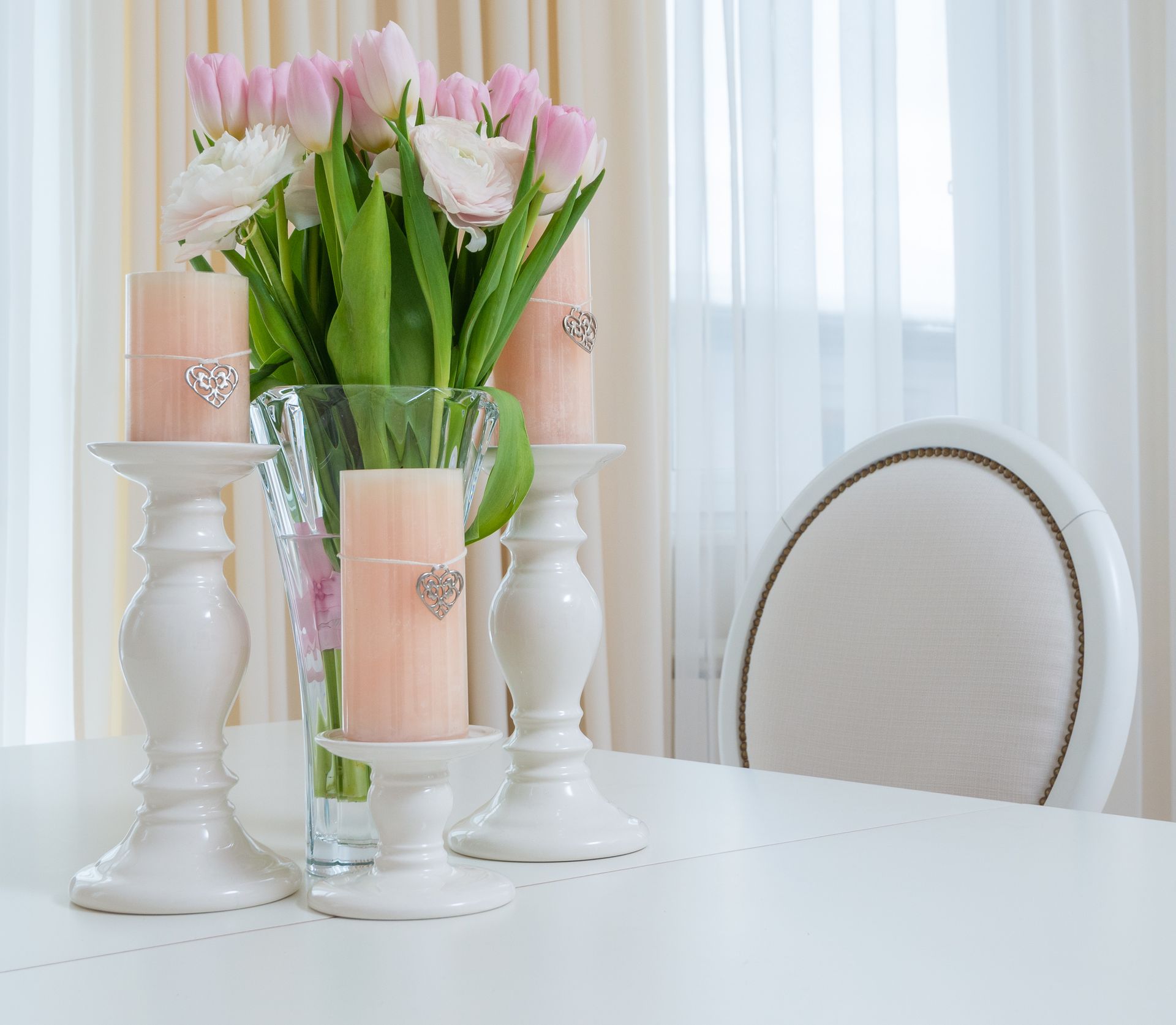 Interior of light dining room with white table with vase with bouquet of flowers and candles in candlesticks near chair next to windows with curtains in daytime