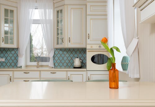 Interior of light empty kitchen with classic cabinets near wooden table with glass vase of tulips flower