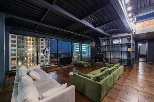 Interior of spacious modern living room with comfortable green and white sofas and panoramic windows in contemporary apartment in evening time