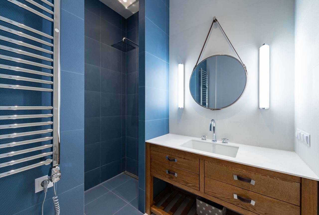Dark blue tiled shower beside a wooden vanity with a round mirror