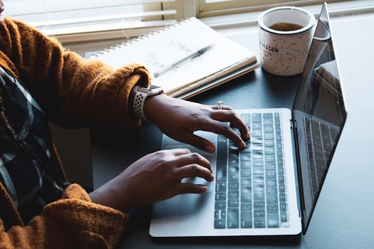 Close-up of a person typing on a laptop indoors with a coffee mug nearby, ideal for remote work themes.