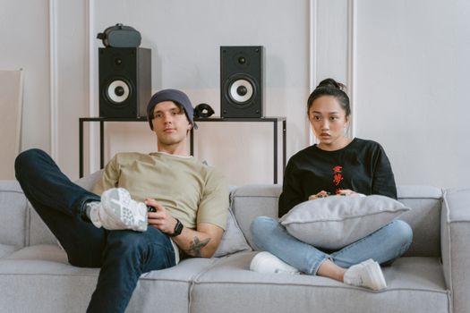 Two young adults enjoying leisure time on a sofa playing video games, surrounded by home speakers.