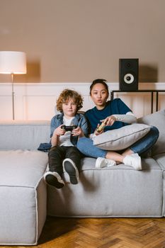 Asian woman and Caucasian boy playing video games together on a cozy couch at home.