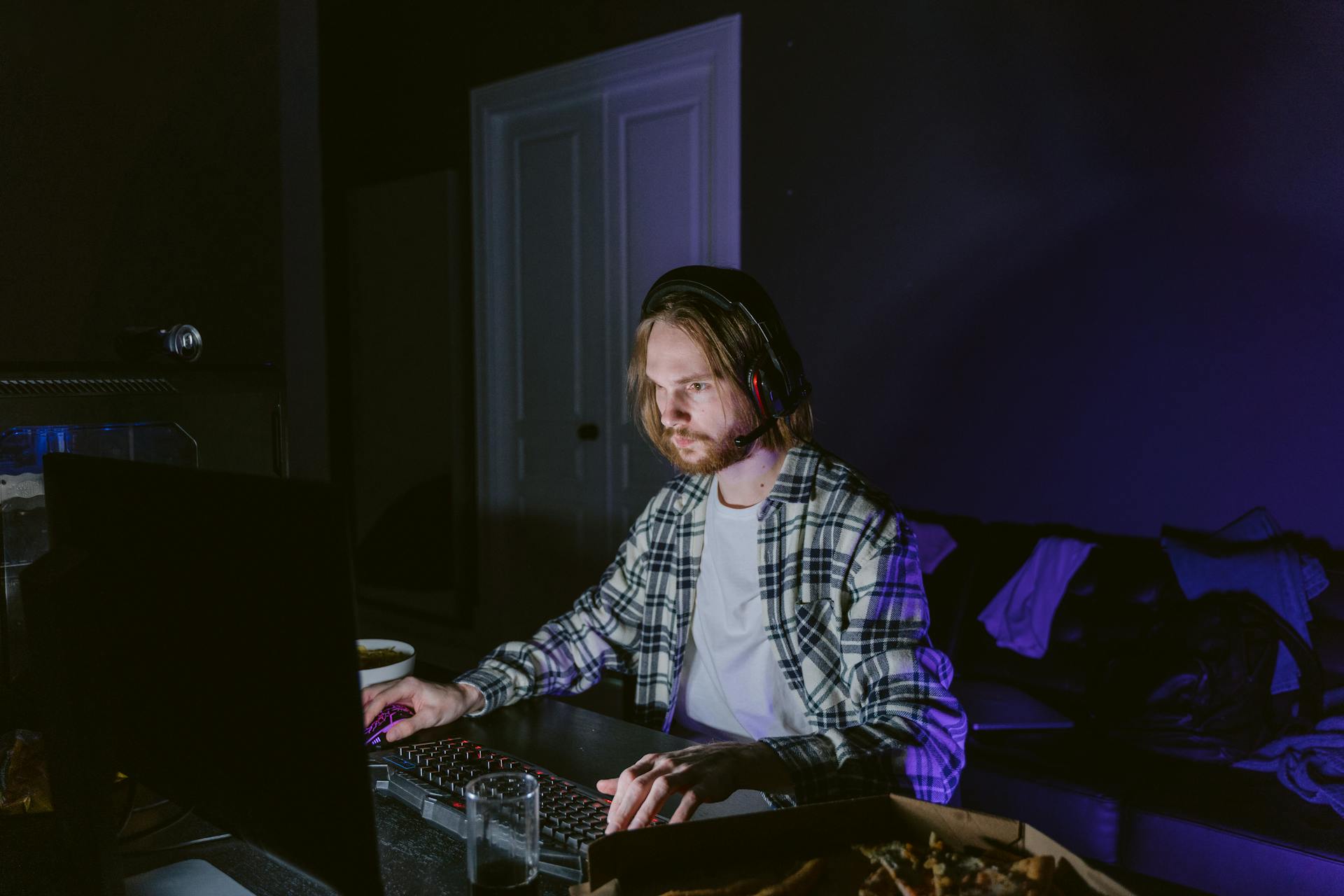 A man wearing headphones plays video games on a computer in a dark room, illuminated by screen light.