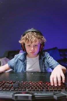 Young boy in denim shirt intensely focused on gaming at computer with glowing keyboard.