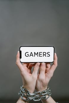 Person's hands in chains holding a smartphone displaying the word 'GAMERS' on a gray background.