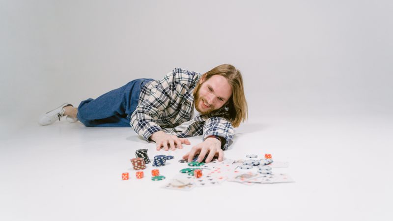 Smiling man with long hair and plaid shirt engaging with casino chips and cards on a white floor.