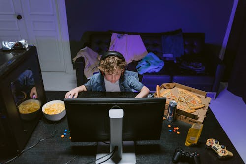 A Boy Wearing a Headset Using a Computer on Table with Food