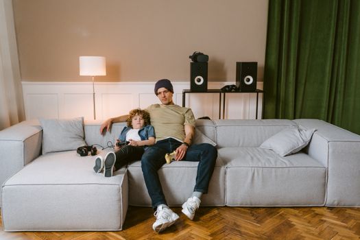 Father and son relaxing on a sofa with game controllers in a cozy living room setting.