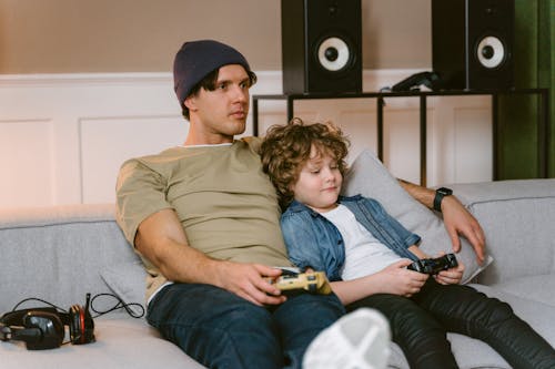 A Man and a Kid Holding Video Game Controllers while Sitting on a Sofa 