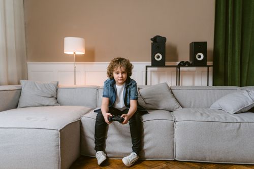 Free Young boy sitting on a comfortable sofa playing video games with a controller indoors. Stock Photo