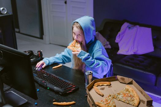 Young girl in blue hoodie eating pizza while gaming at a computer indoors.