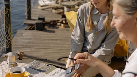 A grandmother and granddaughter enjoying leisure time by a dock with coffee and a newspaper.