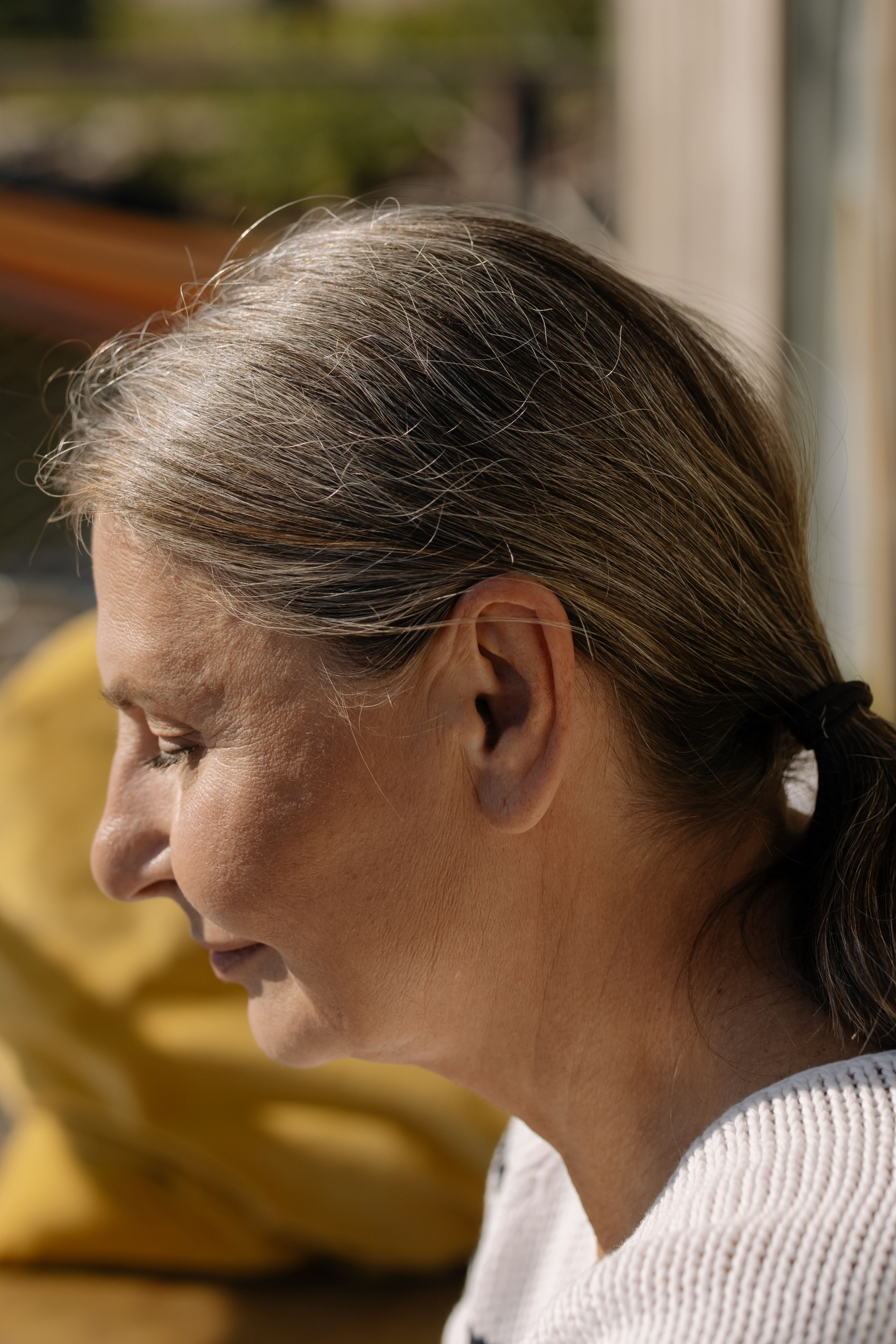Elderly woman with gray hair in a serene profile view, basking in natural sunlight indoors.