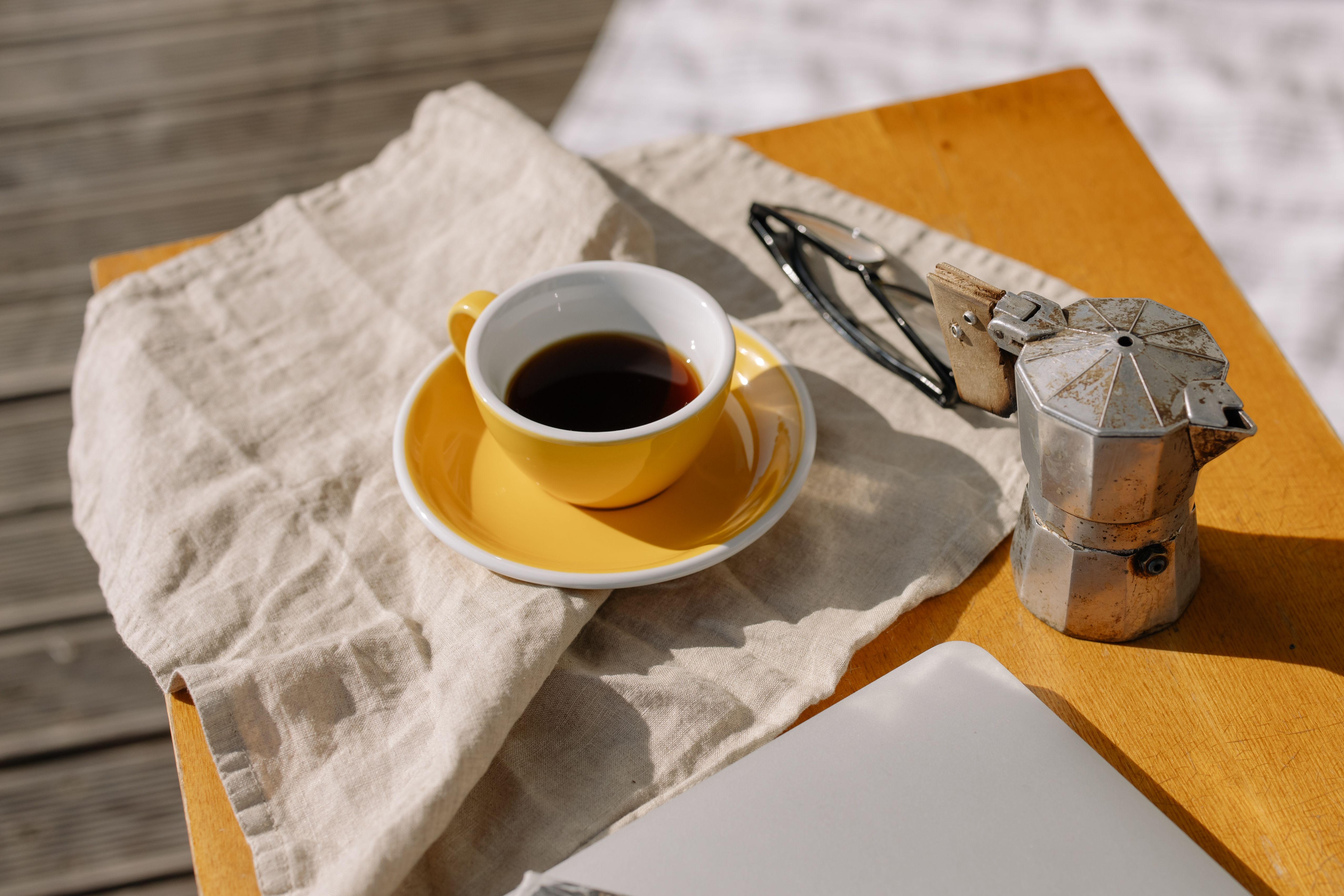 Cozy morning coffee setup with a yellow cup, moka pot, and eyeglasses on a wooden table.