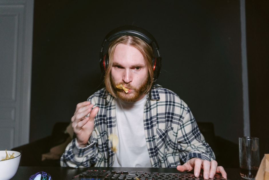 A young man gaming with headphones and snacks, focused and immersed in the activity.