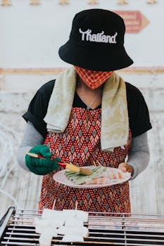 Woman grilling skewers outdoors in Thailand wearing a face mask and bucket hat.