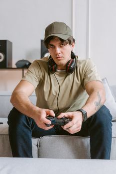 Casual young man gaming on a couch wearing headphones and a cap indoors.