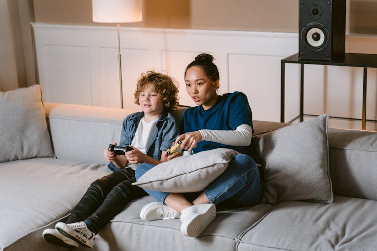 A Woman And A Kid Holding Video Game Controllers While Sitting On A Sofa 