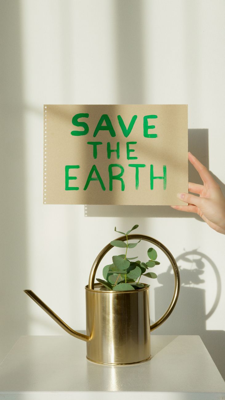 Uma mão segura um cartaz escrito 'SALVE A TERRA' acima de um regador de latão com uma planta de eucalipto sobre um fundo branco.