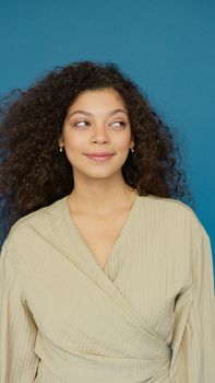 Smiling woman with curly hair wearing a light brown outfit against a blue background.