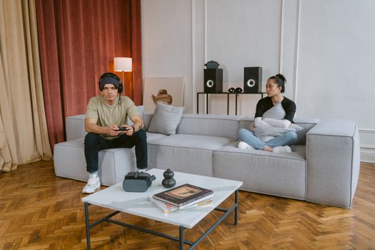 Young interracial couple in stylish living room, one playing video games, the other relaxing.