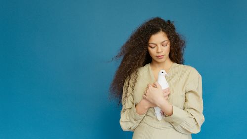 Free Portrait of a young woman in a beige dress holding a white dove against a blue backdrop. Stock Photo