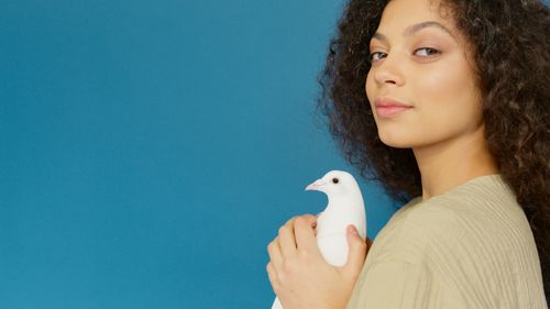 Free A serene portrait of a young woman with curly hair holding a white dove against a blue background. Stock Photo