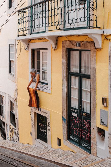 A picturesque street view of an elderly woman hanging a rug outside a Lisbon building with graffiti.