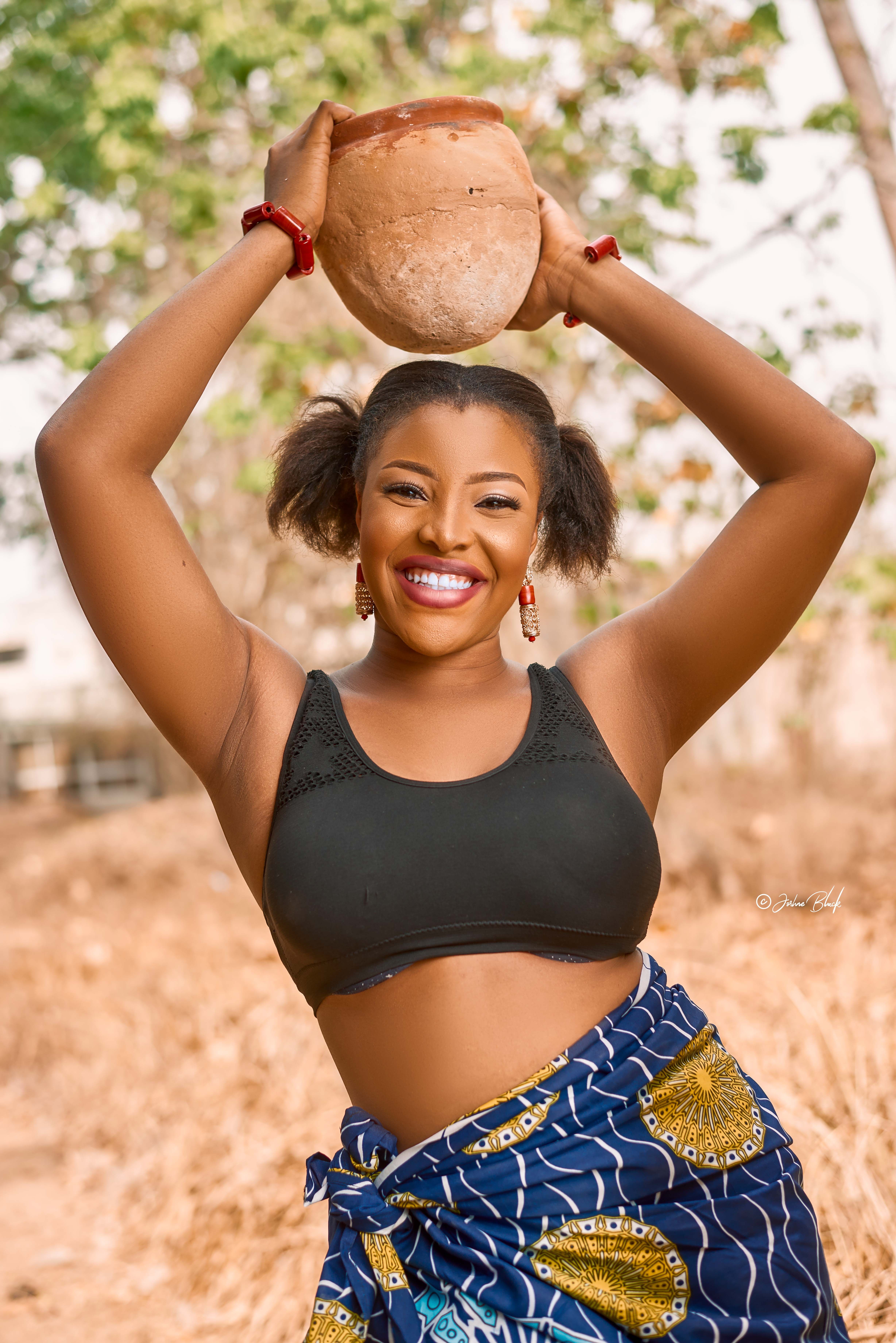 Smiling African American female looking at camera while standing with raised arms with clay pot in hands in suburb area on blurred background