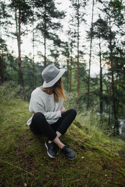 Stylish woman sitting in a serene Russian forest, wearing a knitted sweater and fedora.