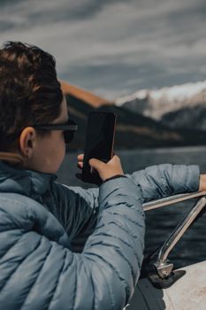 A boy in sunglasses and a puffer jacket captures stunning mountain scenery with his smartphone from a boat.