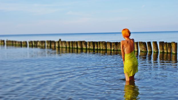 Woman with vibrant red hair standing in calm ocean water near wooden posts.