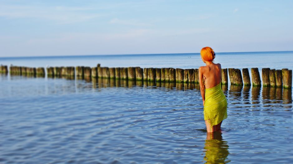 Woman with vibrant red hair standing in calm ocean water near wooden posts.