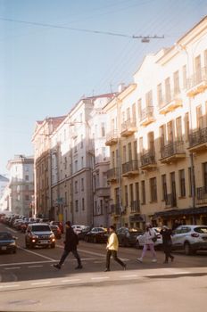 A street view of an urban area with pedestrians crossing and historic buildings under sunlight.