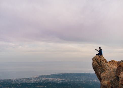 A lone hiker sits on a rocky cliff edge, capturing the scenic city view below.