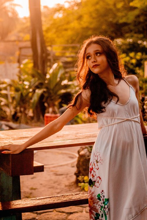 Free A woman in a floral white dress looks away thoughtfully in a warm sunset setting. Stock Photo