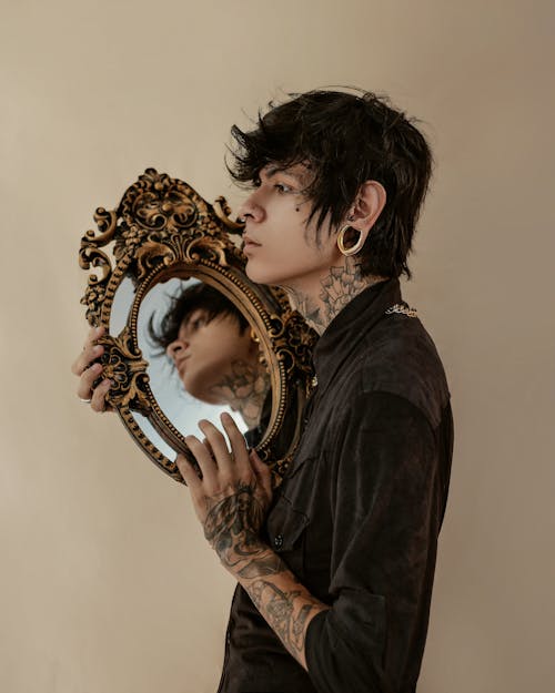 Stylish tattooed man standing with mirror in hand in studio