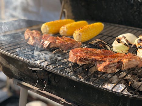Juicy steaks and corn on the cob grilling with smoke rising in a sunny outdoor barbecue.