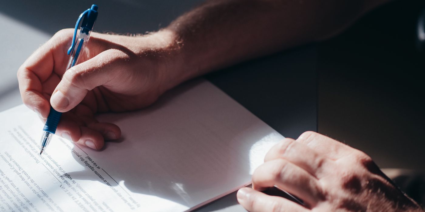 A close-up image of a person's hand signing a document with a blue pen in natural lighting.