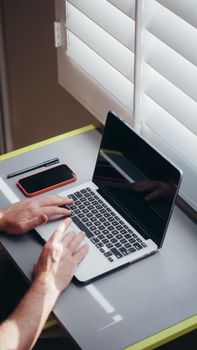 Hands typing on laptop at desk with smartphone and pen, bright window lighting.