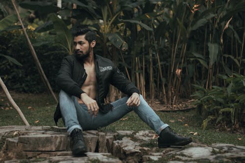 Serious ethnic bearded male with dark hair in trendy clothes sitting on ground in park against green plants and looking away in daytime