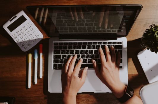 Hands typing on a laptop with a calculator and pens nearby on a wooden table.