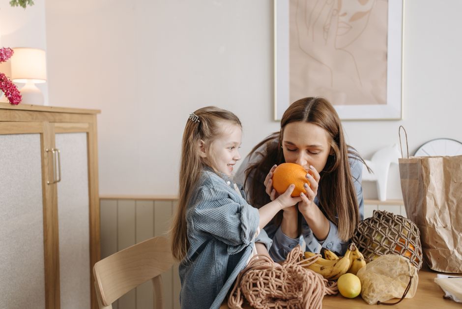 A mother and daughter sharing a moment with fresh oranges at a kitchen table filled with fruit.