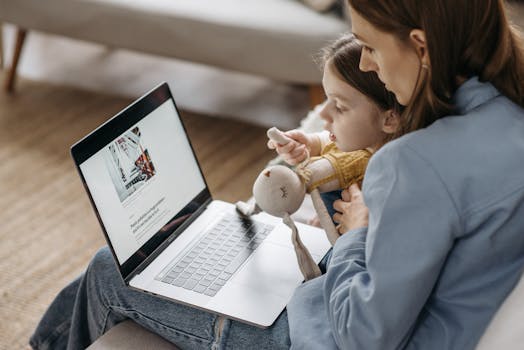 A mother works from home on a laptop with her daughter sitting on her lap, holding a stuffed toy.