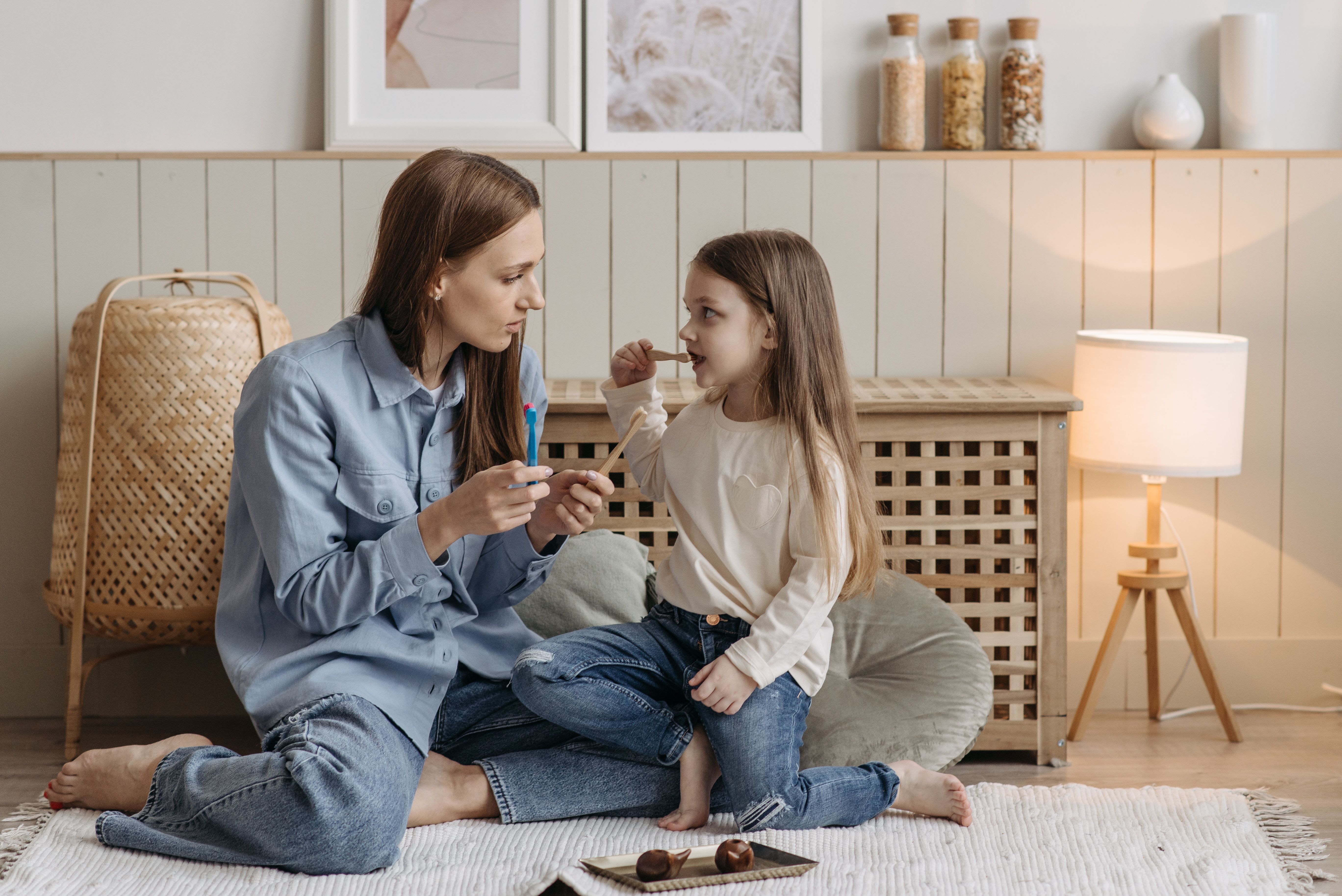 A mother helping her daughter brush teeth while sitting on a cozy carpeted floor.